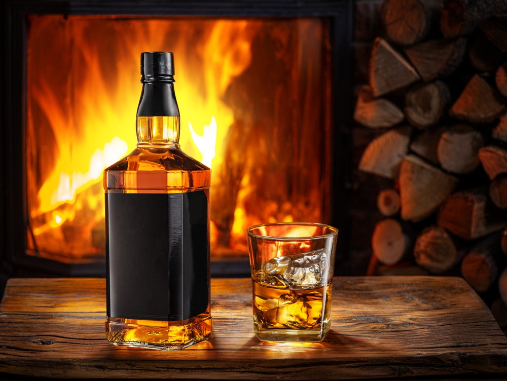 A bottle and glass of whisky on the rocks enjoyed by the fire.