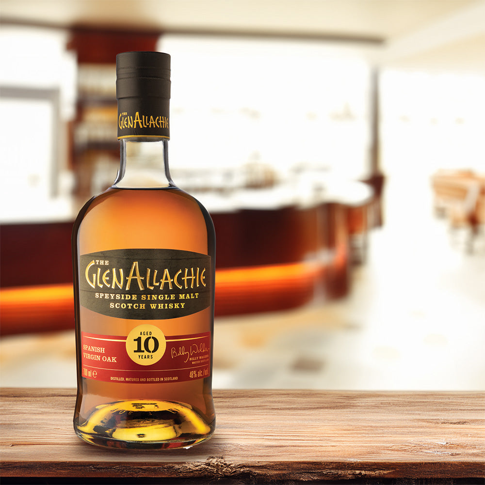 A bottle of GlenAllachie 10 Year Old Spanish Virgin Oak Single Malt Scotch Whisky sits on wood, its black label with gold and red details prominent against a blurred bar. This whisky is matured in Spanish Virgin Oak casks.