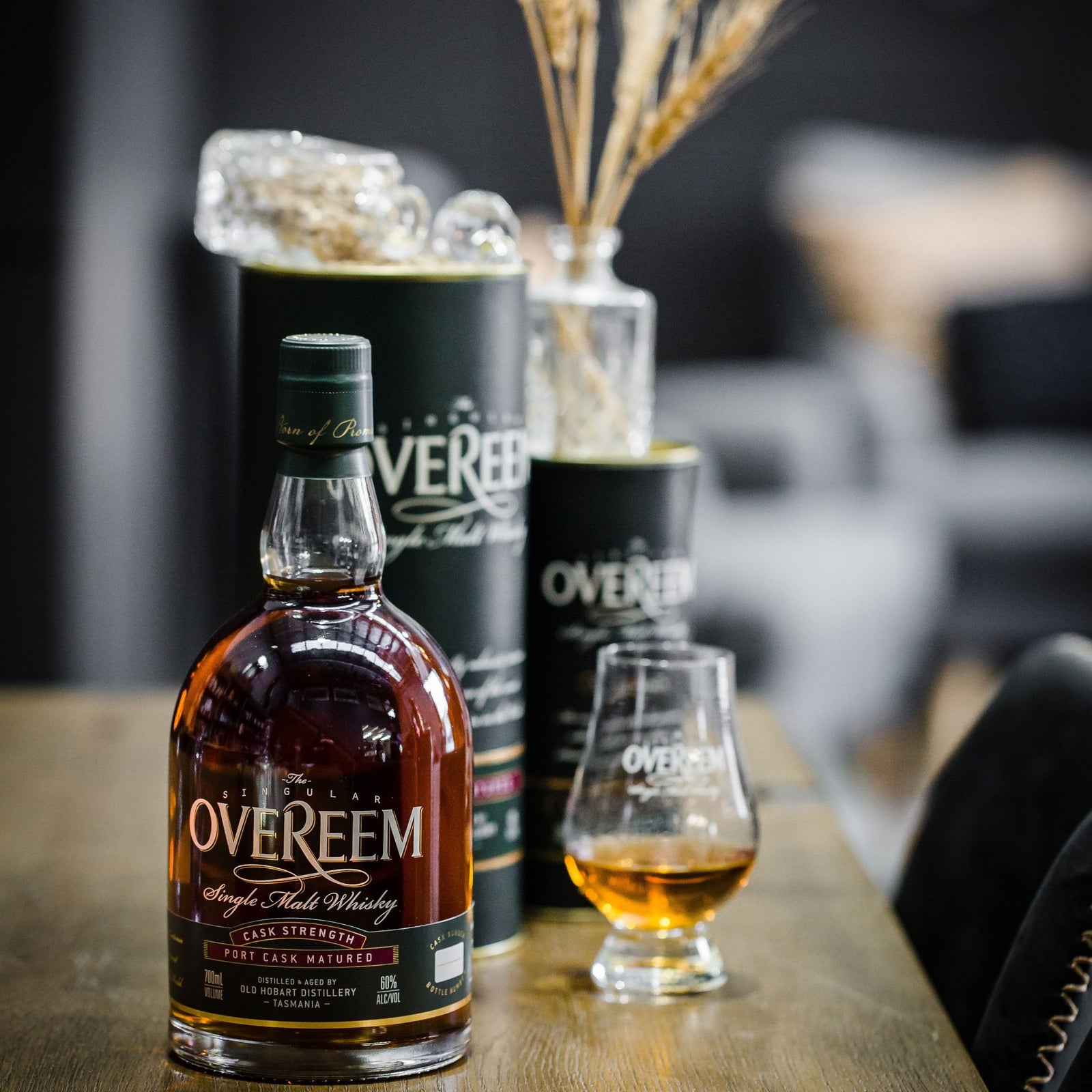 A bottle of Overeem Port Cask Matured Cask Strength Australian Single Malt Whisky rests on a wooden table next to a filled glass, with its cylindrical packaging and decorative dried wheat in the background.