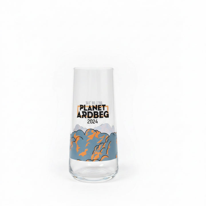 A clear glass tumbler with a printed design reading 'PLANET ARDBEG 2025' and a blue-gray mountain illustration with orange accents, standing on a textured beige carpet against a light wall.