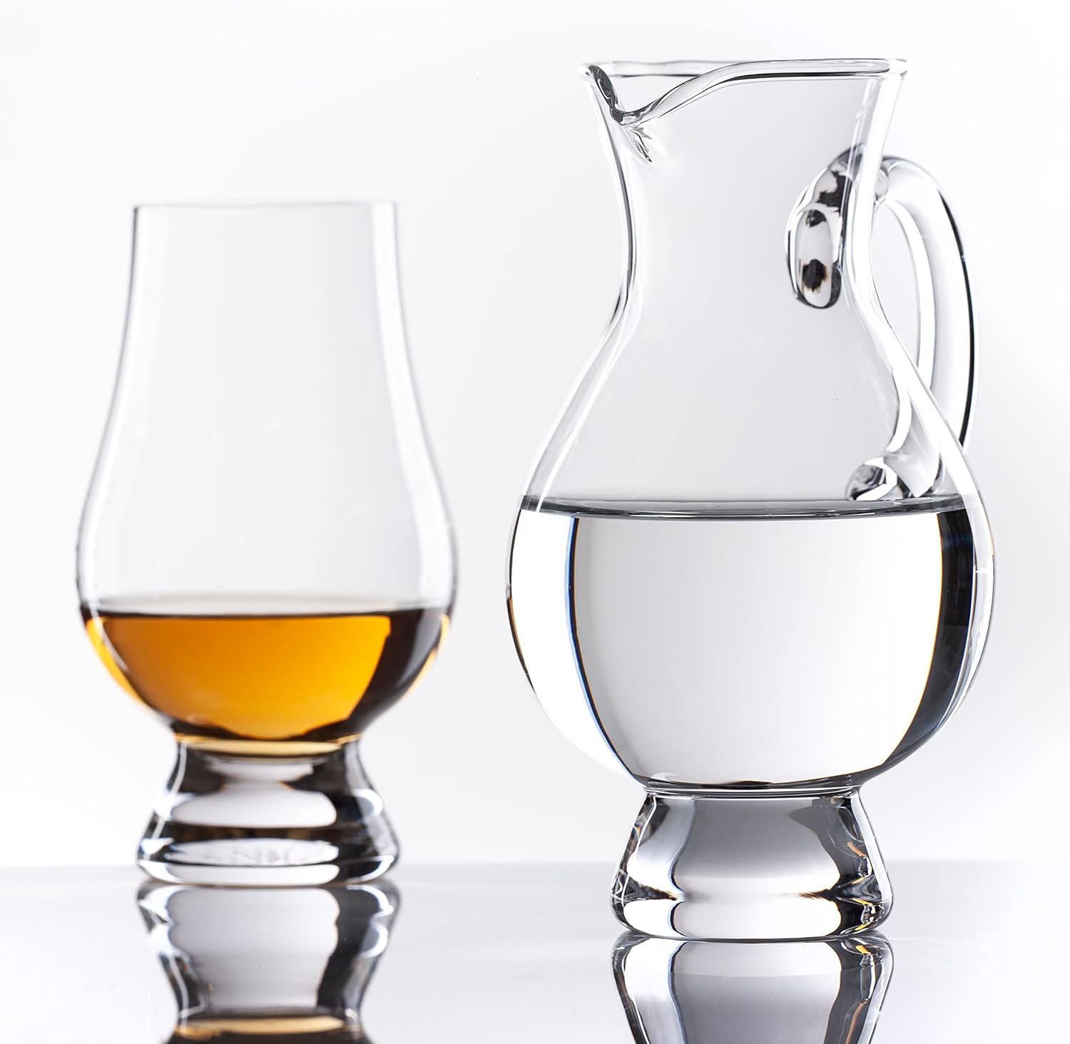 A glass of amber whiskey sits beside a Glencairn Crystal Water Jug filled with water on a reflective surface against a white background—ideal for an elegant whisky tasting.