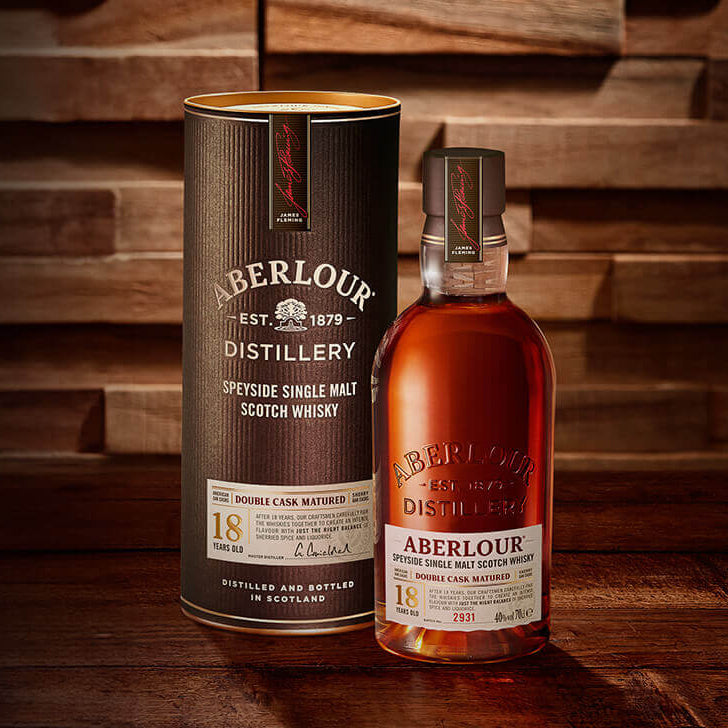 A bottle of Aberlour 18 Year Old Single Malt Scotch Whisky stands next to its cylindrical packaging on a wooden surface, both featuring gold and white labels.