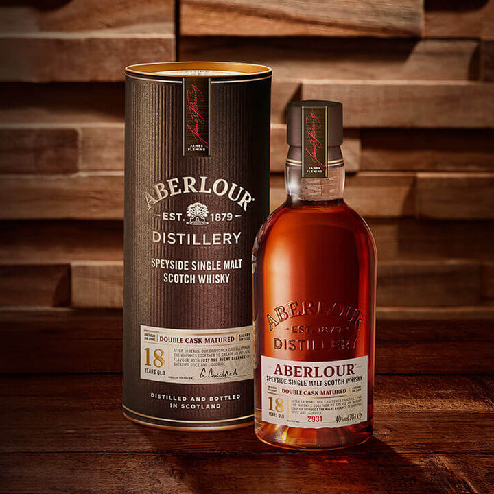 A bottle of Aberlour 18 Year Old Single Malt Scotch Whisky stands next to its cylindrical packaging on a wooden surface, both featuring gold and white labels.