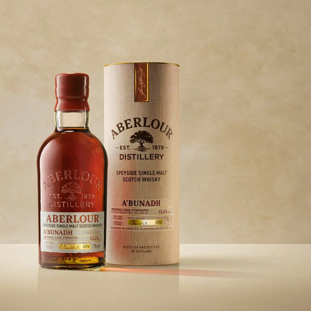 A bottle of Aberlour A’bunadh Single Malt Scotch Whisky stands beside its cylindrical box against a beige background, filled with rich amber whisky and featuring gold and red accents on the label.
