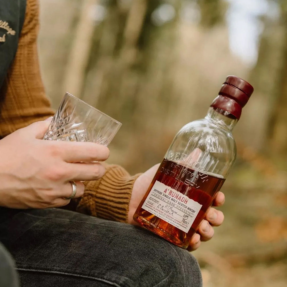 A person holding a glass of cask strength single malt in one hand and a bottle of Aberlour A’bunadh Single Malt Scotch Whisky in the other, sitting outdoors with a blurred forest background.