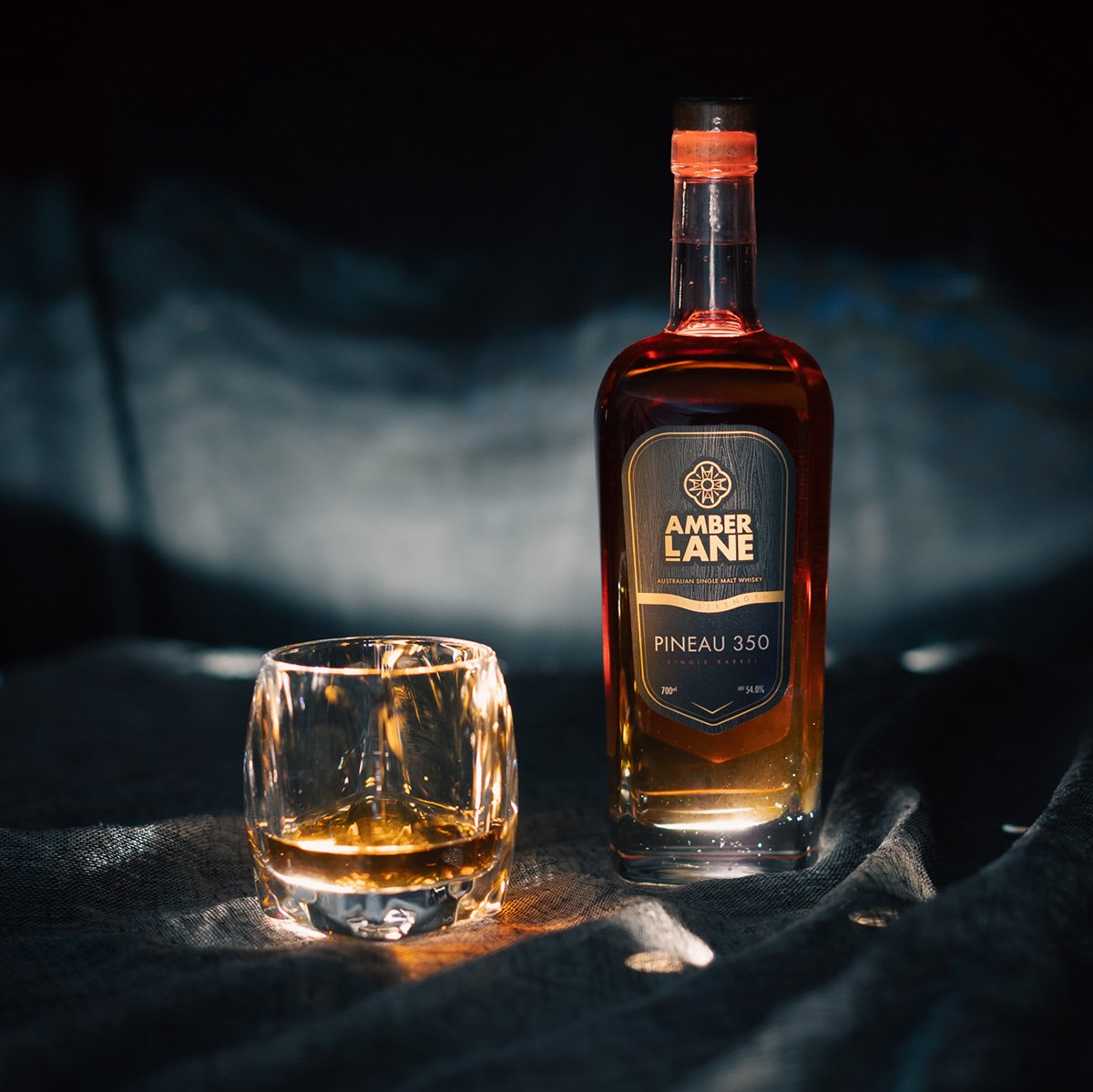 A bottle of Amber Lane Pineau 350 Australian Single Malt Whisky, matured in Pineau des Charentes casks, stands on a dark surface beside a glass of the amber whisky, softly illuminated by moody lighting.