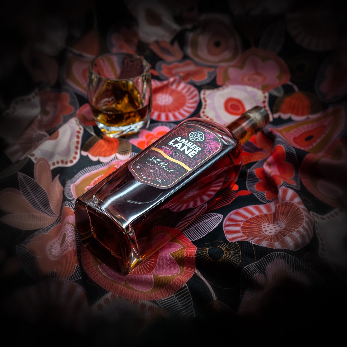 A bottle of Amber Lane Silk Road Australian Single Malt Whisky rests on a colorful, floral-patterned fabric beside a whisky-filled glass, both highlighted against a softly blurred background.