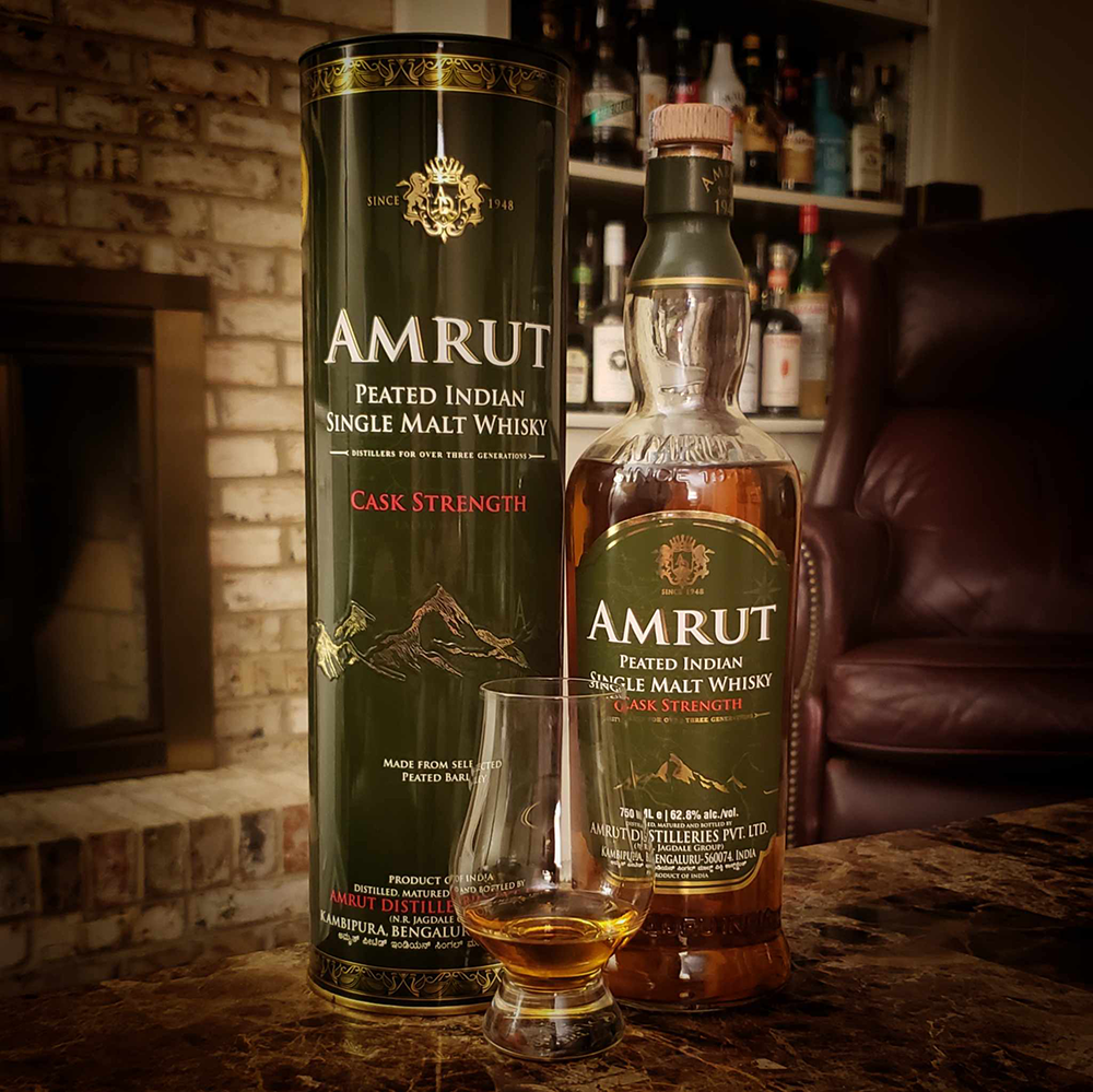 A bottle of Amrut Peated Cask Strength Indian Single Malt Whisky, its cylindrical box, and a filled whisky glass sit on a table in a cozy room with a fireplace and shelves in the background.