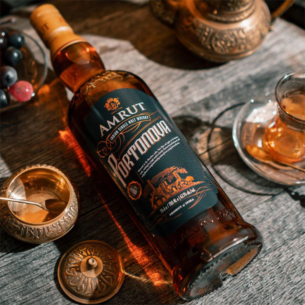 A bottle of Amrut Portonova Indian Single Malt Whisky rests on a wooden table next to a glass of tea, ornate metal cups, and grapes, all bathed in warm light.
