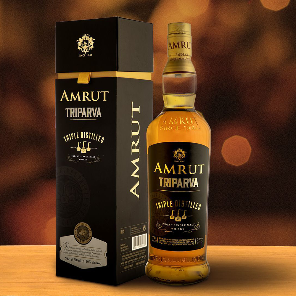 A bottle of Amrut Triparva Indian Single Malt whisky stands next to its elegant black and gold box on a wooden surface, backed by warm, blurred circular lights that highlight the refined smoothness from triple distillation.