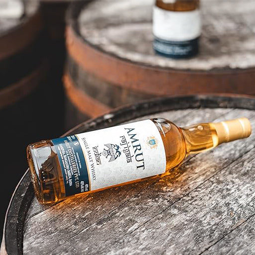 A bottle of Amrut Raj Igala Indian Single Malt Whisky from Amrut Distilleries rests on its side atop a wooden barrel, with another barrel and a blurred bottle in the background.