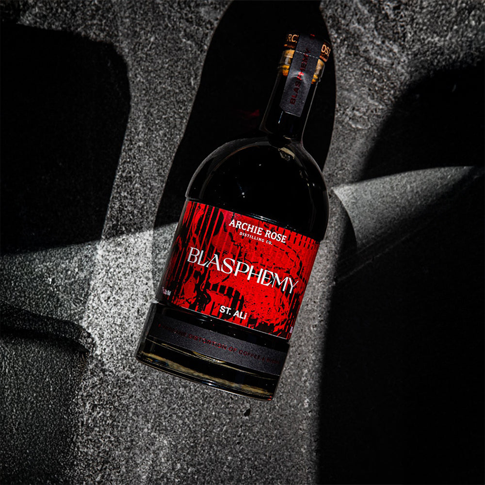 A dark glass bottle labeled Archie Rose x St Ali Blasphemy Coffee Whisky, featuring a striking red and black design, rests on a textured gray surface with deep shadows.