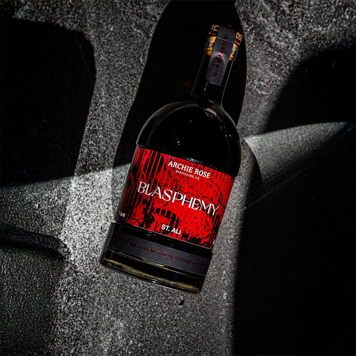 A dark glass bottle labeled Archie Rose x St Ali Blasphemy Coffee Whisky, featuring a striking red and black design, rests on a textured gray surface with deep shadows.