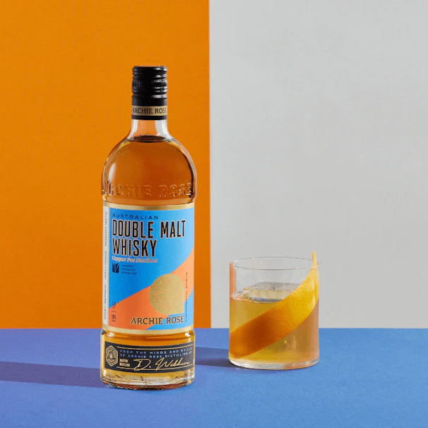 A bottle of Archie Rose Australian Double Malt Whisky sits beside a glass of whisky with an orange peel garnish, set against a split orange, white, and blue background.