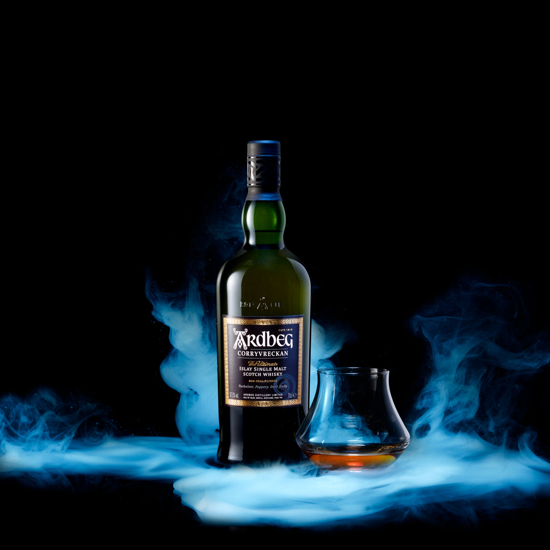 A bottle of Ardbeg Corryvreckan Single Malt Scotch Whisky stands next to a whisky glass holding a small pour, all surrounded by dramatic blue smoke against a black background.