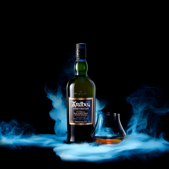 A bottle of Ardbeg Corryvreckan Single Malt Scotch Whisky stands next to a whisky glass holding a small pour, all surrounded by dramatic blue smoke against a black background.