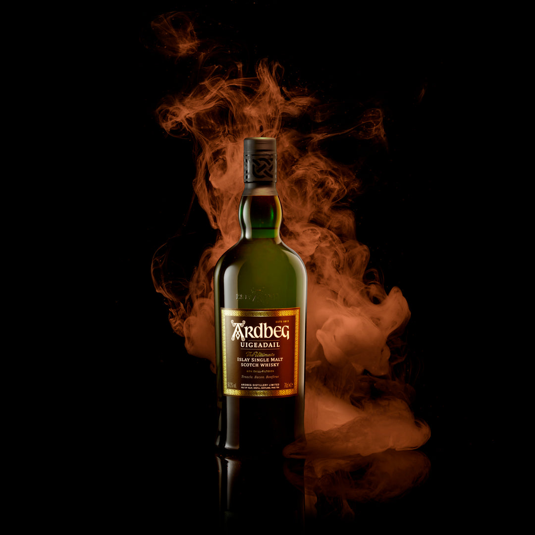 A bottle of Ardbeg Uigeadail Single Malt Scotch Whisky rests on a reflective black surface, framed by swirling orange smoke against a dark backdrop, evoking its deep character and sherry cask influences.