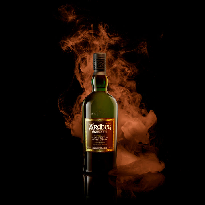 A bottle of Ardbeg Uigeadail Single Malt Scotch Whisky rests on a reflective black surface, framed by swirling orange smoke against a dark backdrop, evoking its deep character and sherry cask influences.