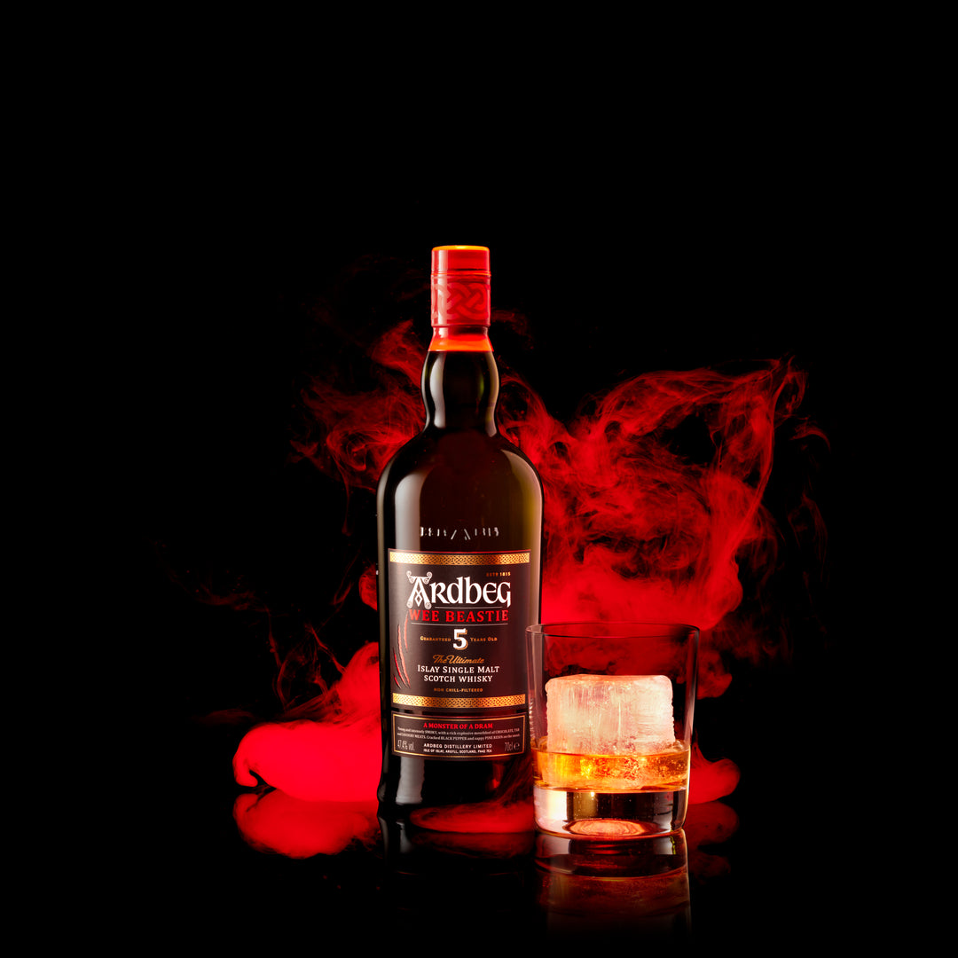 A bottle of Ardbeg 5 Year Old Wee Beastie Single Malt Scotch Whisky stands next to a glass of peated whisky with ice, both surrounded by swirling red smoke against a black background.