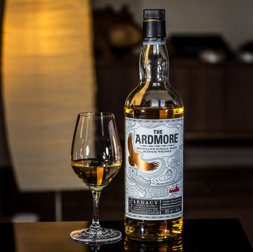 A bottle of Ardmore Legacy Single Malt Scotch Whisky stands by a filled glass on a reflective surface, with a warmly lit background creating a cozy ambiance.