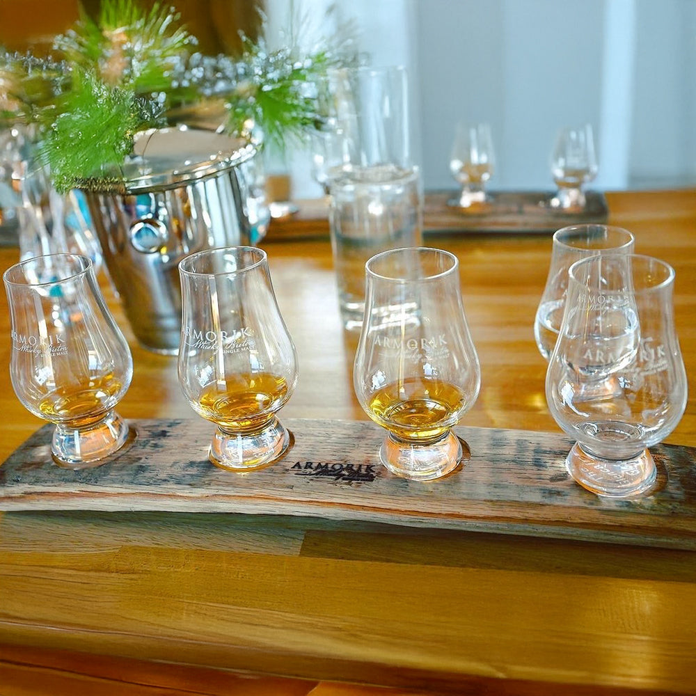 Four whiskey glasses filled with Armorik Pineau des Charentes Single Cask French Whisky sit on a rustic wooden tray atop a polished table, accompanied by a metal ice bucket, glass of water, and vase of greenery for an elegant tasting setup.