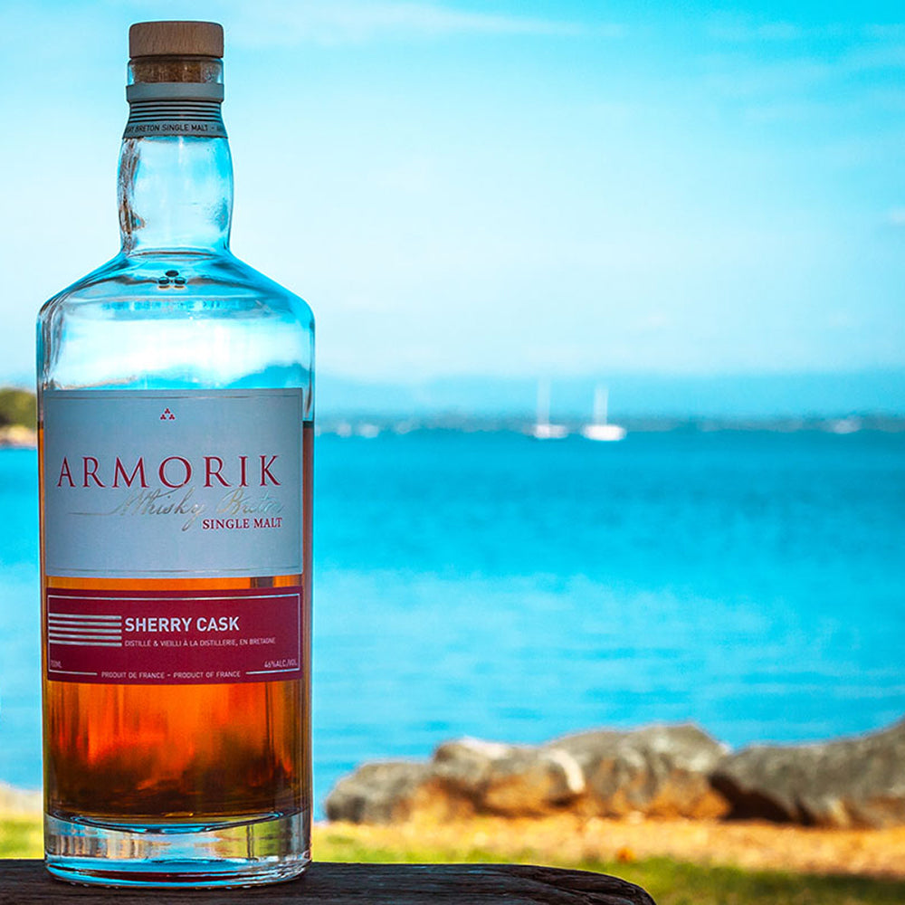 A bottle of Armorik Sherry Cask French Single Malt Whisky rests on a wooden surface outdoors, with blue water, rocks, and blurred sailboats in the background under a clear sky.