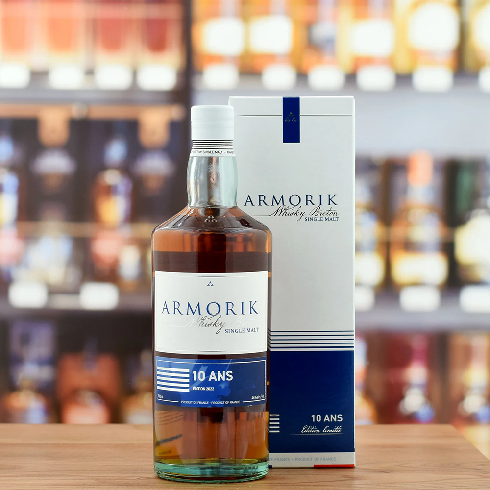 A bottle of Armorik 10 Ans (10 Year Old) French Single Malt Whisky sits on a wooden surface next to its matching white and blue box, with blurred shelves of bottles in the background.