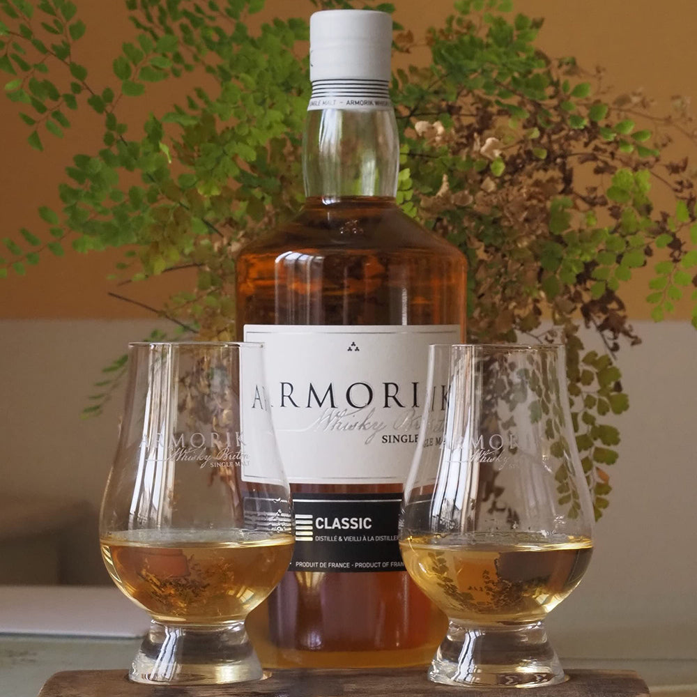 A bottle of Armorik Classic French Single Malt Whisky stands behind two filled glasses on a wooden surface, with green leafy plants in the background.