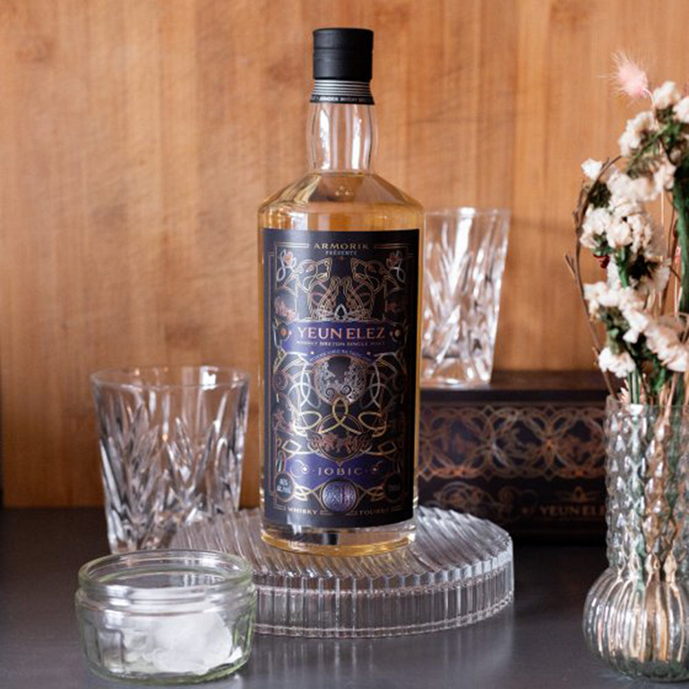 A bottle of Armorik Yeun Elez Jobic French Single Malt Whisky is displayed on a ribbed glass tray with two crystal glasses, a jar of ice, and a floral arrangement, all set against a wooden background.