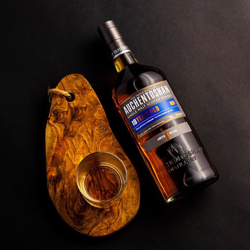 A bottle of Auchentoshan 18 Year Old Single Malt Scotch Whisky sits beside a glass of whisky on a wooden board, set against a black background.