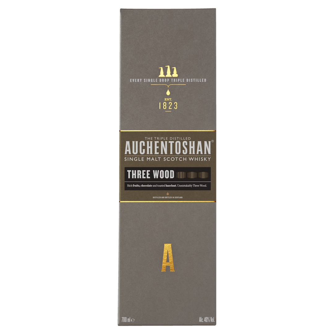 A box of Auchentoshan Three Wood Single Malt Scotch Whisky features gold and white text on a gray background, prominently displaying the brand logo, whisky details, and highlighting its triple maturation process.