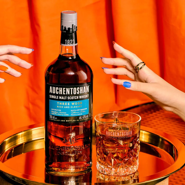 A bottle of Auchentoshan Three Wood Single Malt Scotch Whisky and a whisky glass rest on a gold tray, as two manicured hands reach for the triple-matured bottle set against an orange curtain backdrop.