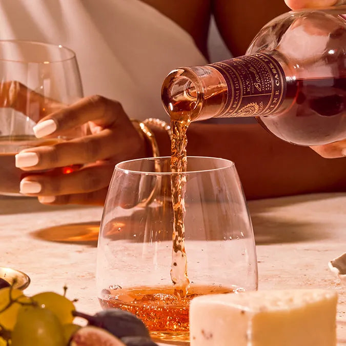 A hand pours amber Basil Hayden Red Wine Cask Finish Bourbon into a glass on a table with cheese, fruit, and another guest enjoying a drink—capturing an elegant, social moment perfect for savoring this unique bourbon.