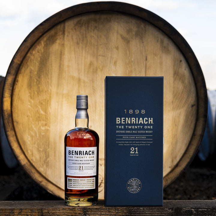 A bottle of Benriach The Twenty One Single Malt Scotch Whisky and its navy box are displayed outdoors before a large wooden barrel, with blurred barrels and a bright sky in the background.