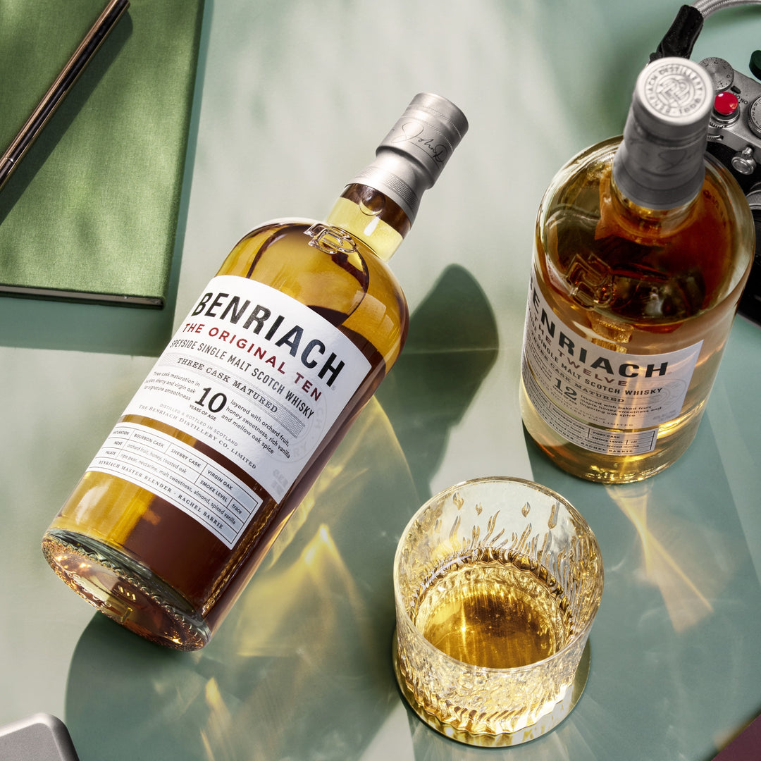 Two bottles of Benriach whisky, including Benriach The Original Ten Single Malt Scotch Whisky, sit on a glass table beside a whisky tumbler. A controller and a green notebook are also on the table.