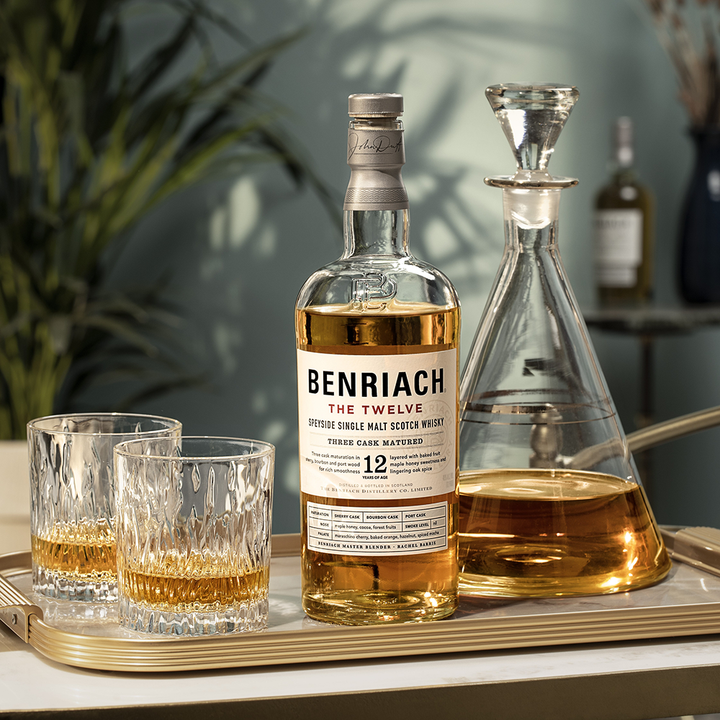 A bottle of Benriach The Smoky Twelve Single Malt Scotch Whisky is displayed on a tray with two filled glasses and a decanter, while a blurred bottle and plant appear in the background of a brightly lit room.