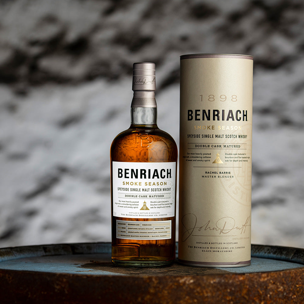 A bottle of Benriach Smoke Season Single Malt Scotch Whisky stands beside its cylindrical box on a wooden barrel, emphasizing this bold Speyside whisky against a muted gray and white background.