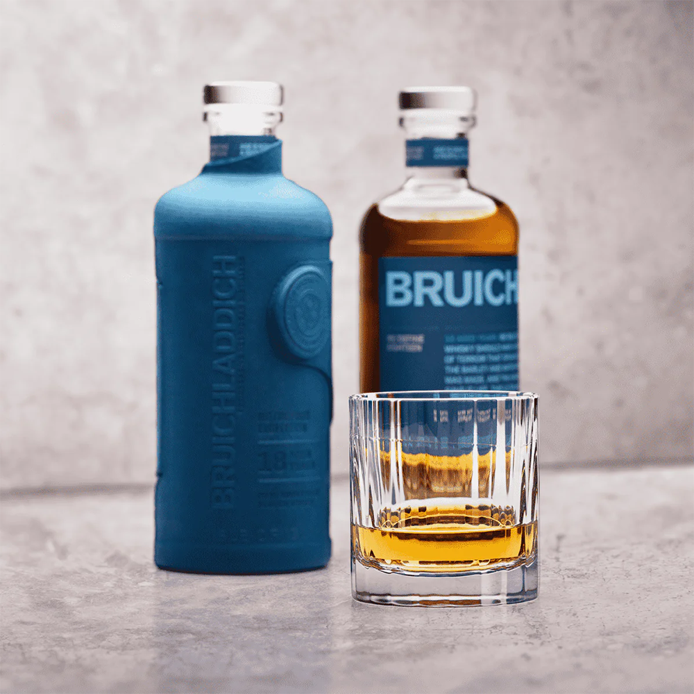 Two bottles of Bruichladdich 18 Year Old Single Malt Scotch Whisky—one blue and one transparent with amber liquid—stand behind a clear glass on a light gray surface against a neutral background.