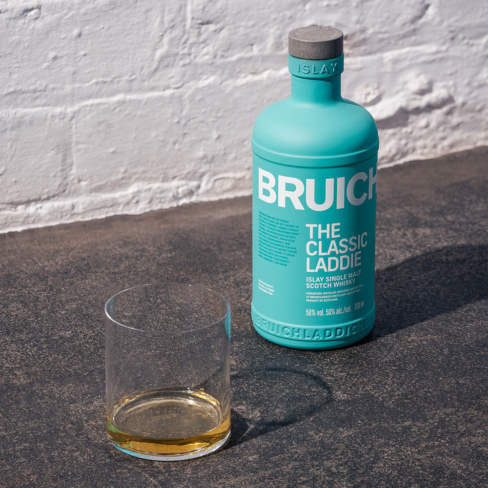 A turquoise bottle of Bruichladdich The Classic Laddie Single Malt Scotch Whisky stands on a sunlit surface beside a clear glass with a small pour, with a white brick wall in the background.