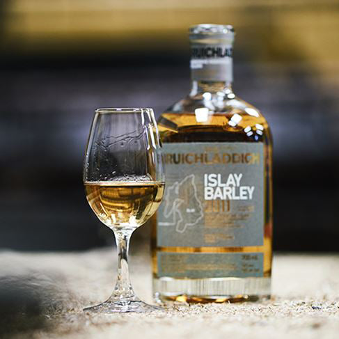 A bottle of Bruichladdich Islay Barley Single Malt Scotch Whisky stands beside a filled whisky glass on a textured surface, with a softly blurred background.