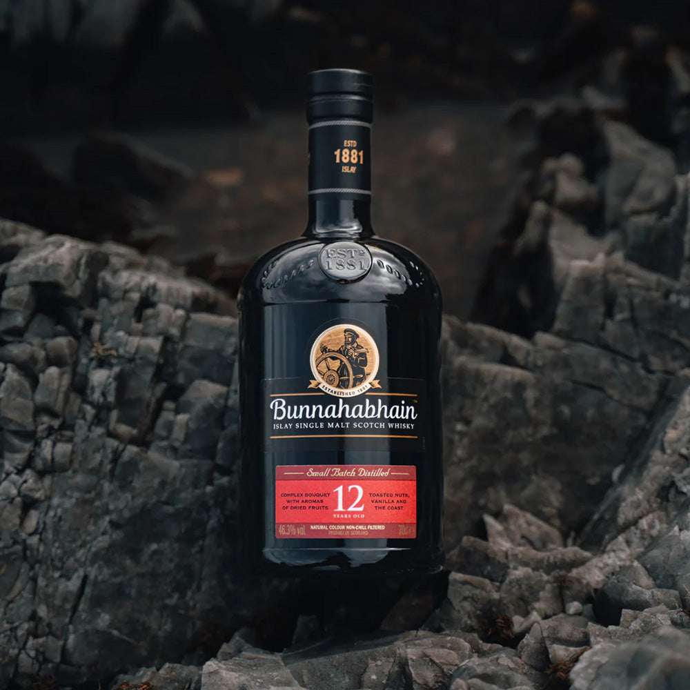 A bottle of Bunnahabhain 12 Year Old Single Malt Scotch Whisky sits on dark, rugged rocks, its label and branding clearly visible.