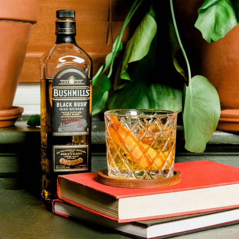 A bottle of Bushmills Black Bush Irish Whiskey sits next to a glass garnished with an orange twist atop a red book and coaster, set against green potted plants, showcasing its signature rich sherry cask influence.