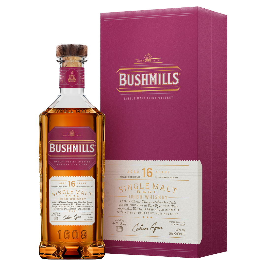 A bottle of Bushmills 16 Year Old Single Malt Irish Whiskey with a cork top stands beside its matching maroon box, both displaying gold and cream label details and product information.