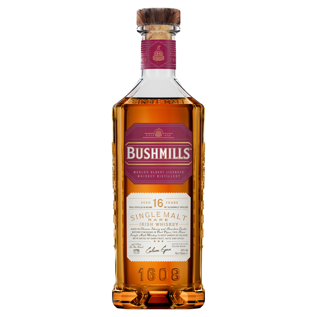 A bottle of Bushmills 16 Year Old Single Malt Irish Whiskey, featuring a hexagonal shape, amber liquid, red and gold label, and cork stopper, is shown against a plain white background.