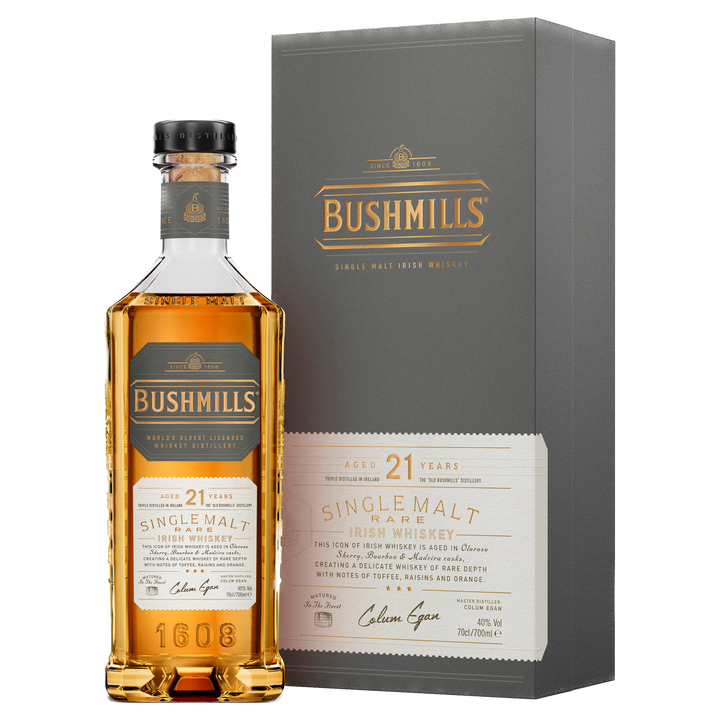 A bottle of Bushmills 21 Year Old Single Malt Irish Whiskey is displayed in front of its gray and gold gift box, both labels showcasing this exceptional single malt and revealing the bottle’s rich amber color.