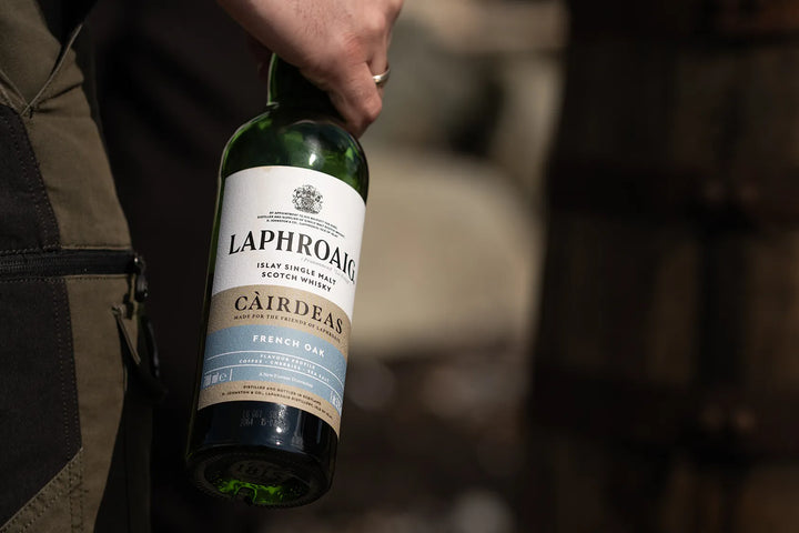 A hand holding a bottle of Laphroaig Cairdeas French Oak single malt Scotch whisky against a dark, blurred background.