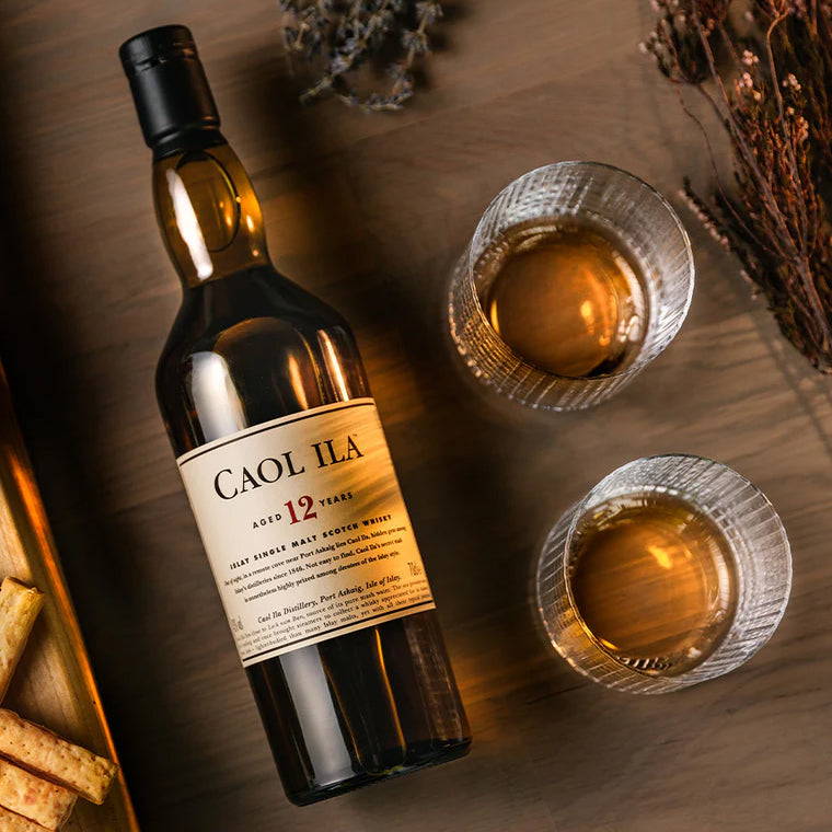A bottle of Caol Ila 12 Year Old Single Malt Scotch Whisky sits on a wooden surface next to two glasses of peated whisky, assorted crackers, and scattered dried herbs.