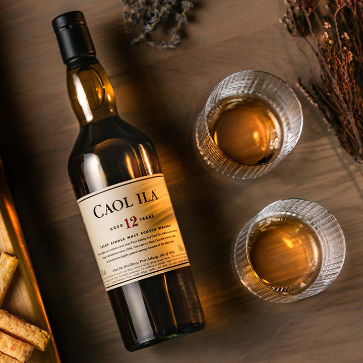 A bottle of Caol Ila 12 Year Old Single Malt Scotch Whisky sits on a wooden surface next to two glasses of peated whisky, assorted crackers, and scattered dried herbs.