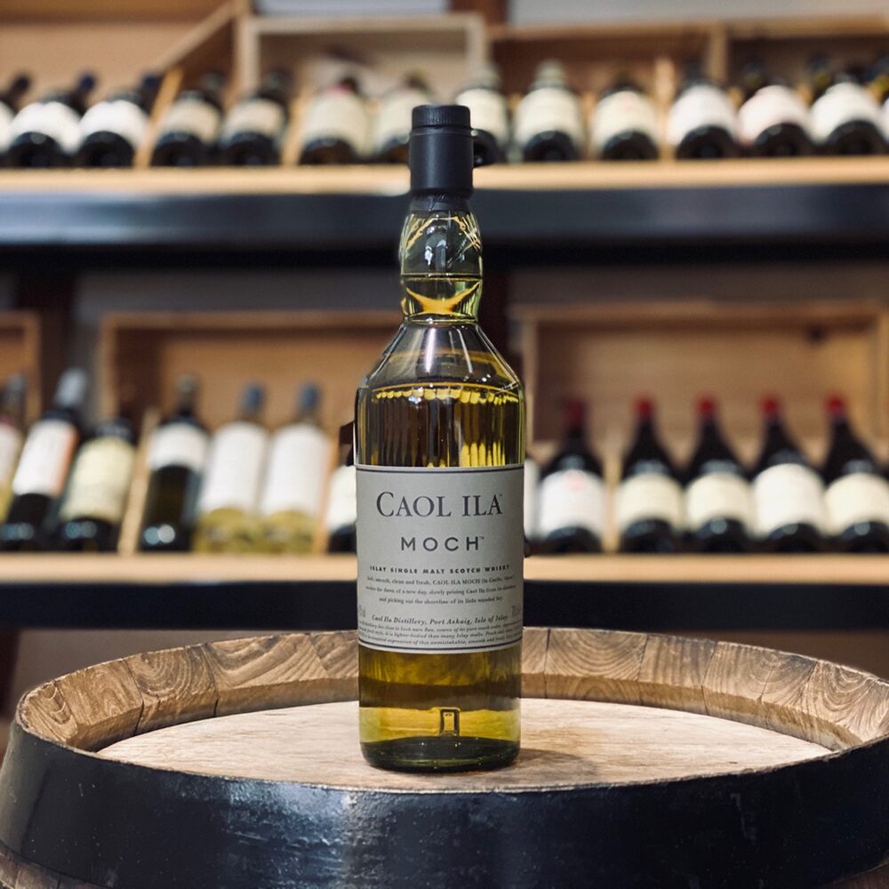 A bottle of Caol Ila Moch Single Malt Scotch Whisky stands on a wooden barrel, with blurred shelves of assorted wine bottles in the background.
