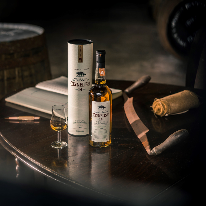 A bottle of Clynelish 14 Year Old Single Malt Whisky is displayed with its box, a filled glass, an open notebook, a pen, and vintage tools on a dark wooden table in soft lighting.